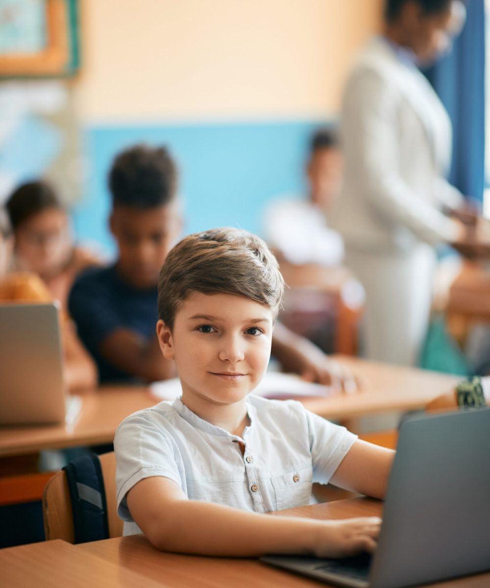 Smiling elementary student using laptop while e-learning during class in the classroom and looking at camera.